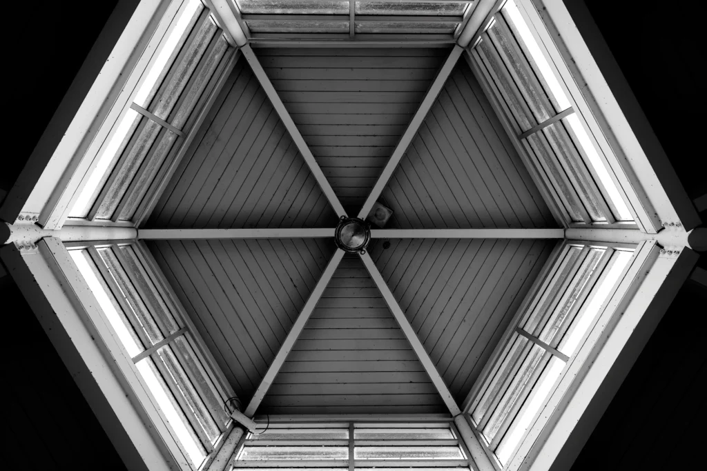 Black and white photo. A hexagonal structure of brightly lit panes on the sides, and crossbeams in the middle.