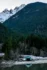 Color photo. At the bottom, a pale green boathouse sits above the waters of Lake Diablo, in which it is reflected. The beach is covered in a dusting of snow. Bare hardwood trees line the near shoreline, and beyond them, rising rapidly upward, and dark slopes of pine. Snow covered peaks, with dark rocks, are visible in the upper left of the frame.