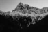 Black and white photo. Pyramid Peak, in the North Cascades National Park It is snow covered, with dark rocks peaking through showing the sheer faces of the cliffs below the peak. Below it is a tree-filled snowscape. An angle or valley formed by two dark tree-lined slopes on either side at the bottom of the frame creates an angle in the snowscape that mirrors the mountain peak above.