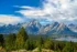 Color photo. The Grand Tetons in the middle frame, from a distance of a few miles. Their peaks are craggy and pointed, with patches of snow in the deeper crevasses. In the immediate foreground there is a line of pine trees. In the valley between is a green forest, with a lake visible on the right. The deep blue sky is filled with puffy white clouds, some of which are streaked, indicating rain.