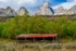 Color photo. A building, possibly a barn made of wooden logs, with a wooden hitching rail in front, in a field of green and sage grass. Its roof is red. Behind it is a line of trees with green leaves, tinging yellow towards autumn. Looming above them all are the jagged peaks of the Tetons in a cloud filled sky.