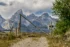 Color photo. A gate of wooden logs, between two wooden posts. The left side of the gate is taller than the right, and a log goes diagonally from the top left to the lower right. Below the gate is a narrow dirt road, and a green tree is on the right. Beyond the gate the jagged peaks of the Tetons rise up into a cloud filled sky.