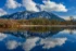 Color photo. Mount Si rises above the waters of Borst Lake. The far shore is lined with bare trees. The sheer faces of the mountain are white with new snow. Three puffy white clouds are in the blue sky above the mountain. The top half of the photo is reflected in the water of the bottom half.