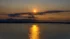 Color photo. A setting sun above the Olympic Mountains, which are seen in silhouette. Between the sun and the mountains are clouds, but elsewhere the sky is clear and golden. The bottom half of the photo are the waters of Puget Sound, upon which the sunlight is casting a lane of bright reflection. A line of sailboats are crossing the lane.
