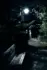 Color photo. A wooden bench next to a paved path. It is night, and there is a bright lamppost visible through a gap in the trees around the bench and path. The bench catches the light, but the trees and ground around it are in deep shadow. The photo is in color, but the colors are muted.