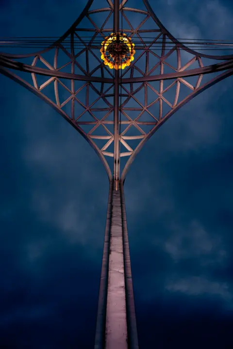 Color photo. A tall pillar rises up from the bottom of the frame and rises to meet a lattice at the top. A ring of golden light is present near the top as well.